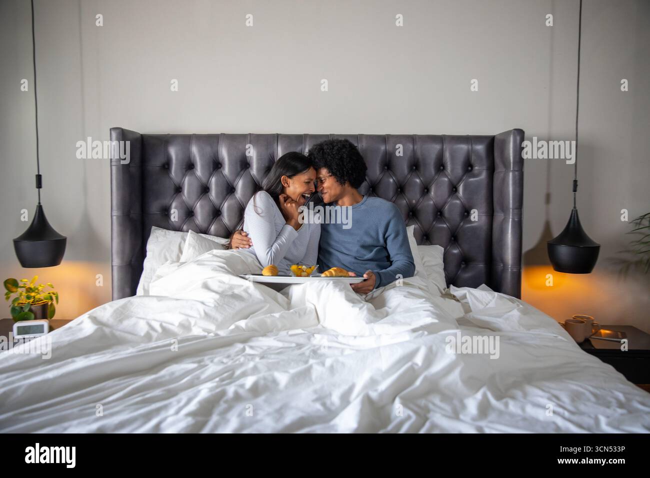 Couple diversifié partageant plateau de petit déjeuner en bois avec jus d'orange, croissants dans la chambre principale Banque D'Images Couple diversifié partageant plateau de petit déjeuner en bois avec jus d'orange, croissants dans la chambre principale Banque D'Images