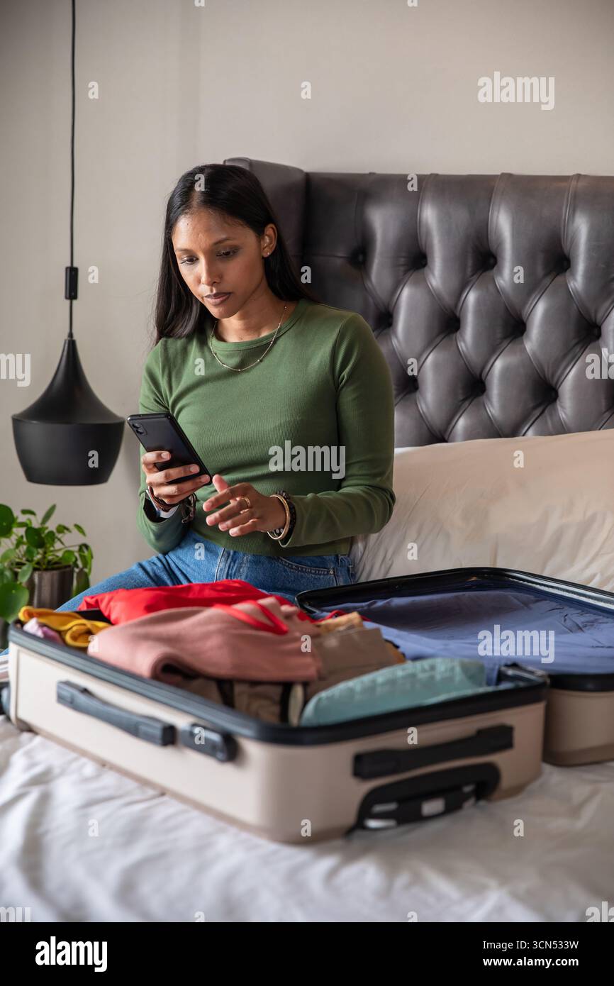 Femme indienne emballant une valise ouverte sur le lit tout en utilisant un smartphone dans la chambre, tête de lit touffetée Banque D'Images Femme indienne emballant une valise ouverte sur le lit tout en utilisant un smartphone dans la chambre, tête de lit touffetée Banque D'Images
