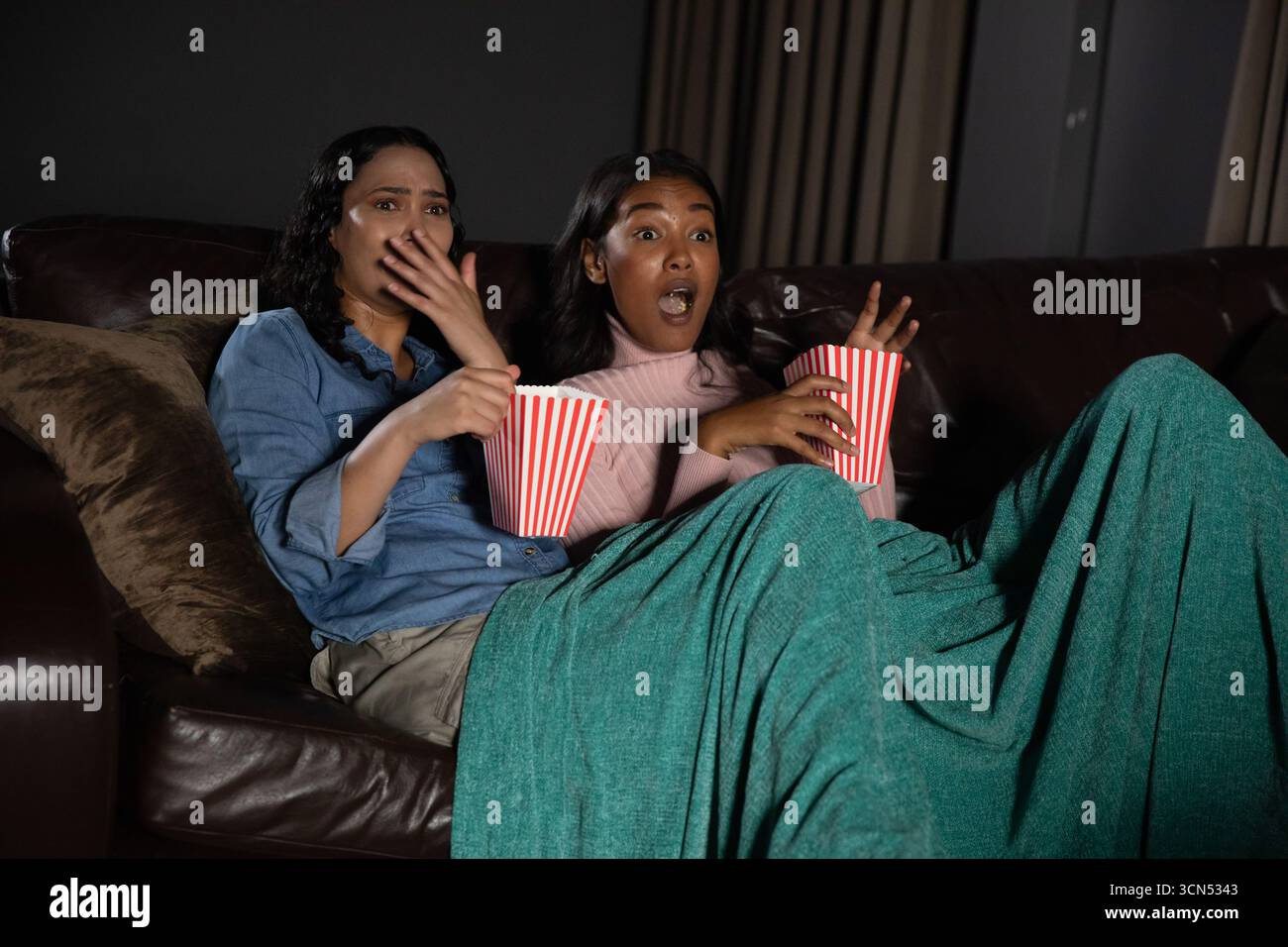 Diverses amies féminines réagissant avec surprise, regardant le film sur le canapé du salon avec pop-corn Banque D'Images Diverses amies féminines réagissant avec surprise, regardant le film sur le canapé du salon avec pop-corn Banque D'Images
