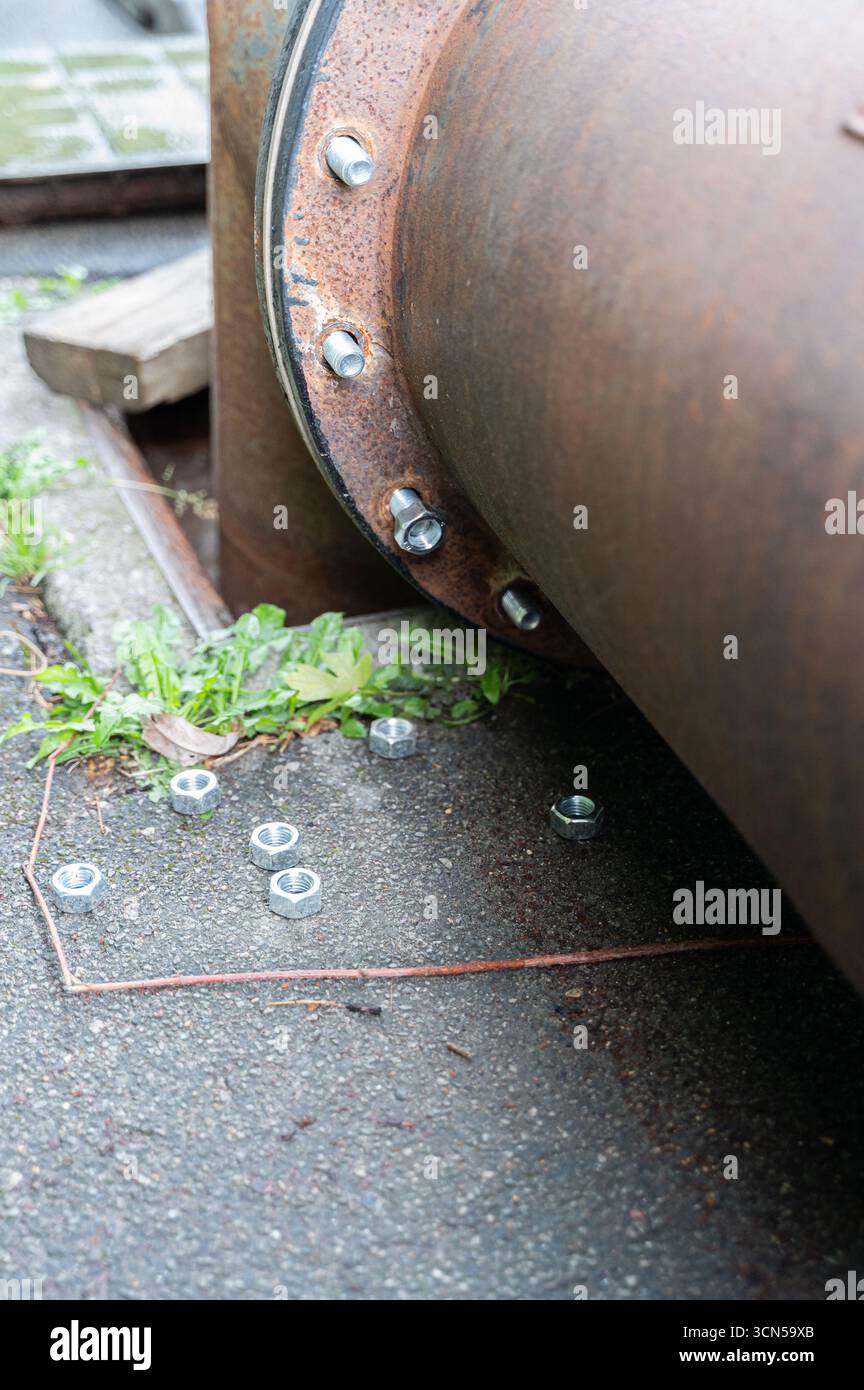 Une vue rapprochée d'un tuyau industriel rouillé sur béton, avec des écrous métalliques et la verdure environnante. Banque D'Images