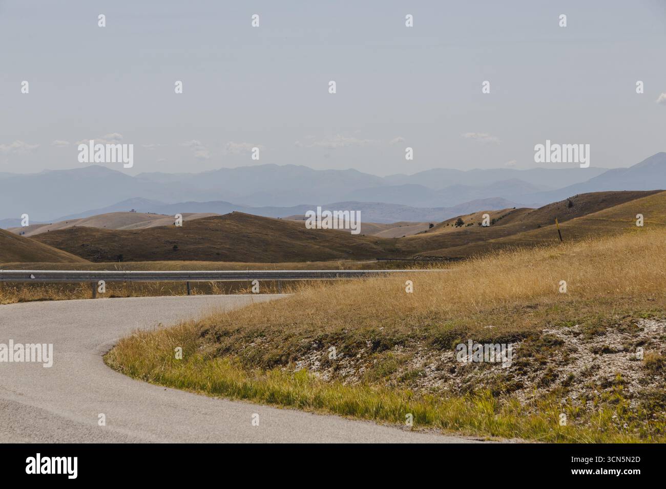 Vue sur la route sinueuse coupe à travers les prairies dorées ondulantes, menant vers les montagnes bleues brumeuses, un paysage serein, Isola del Gran Sasso D'italia, Abruzzes, Italie. Banque D'Images