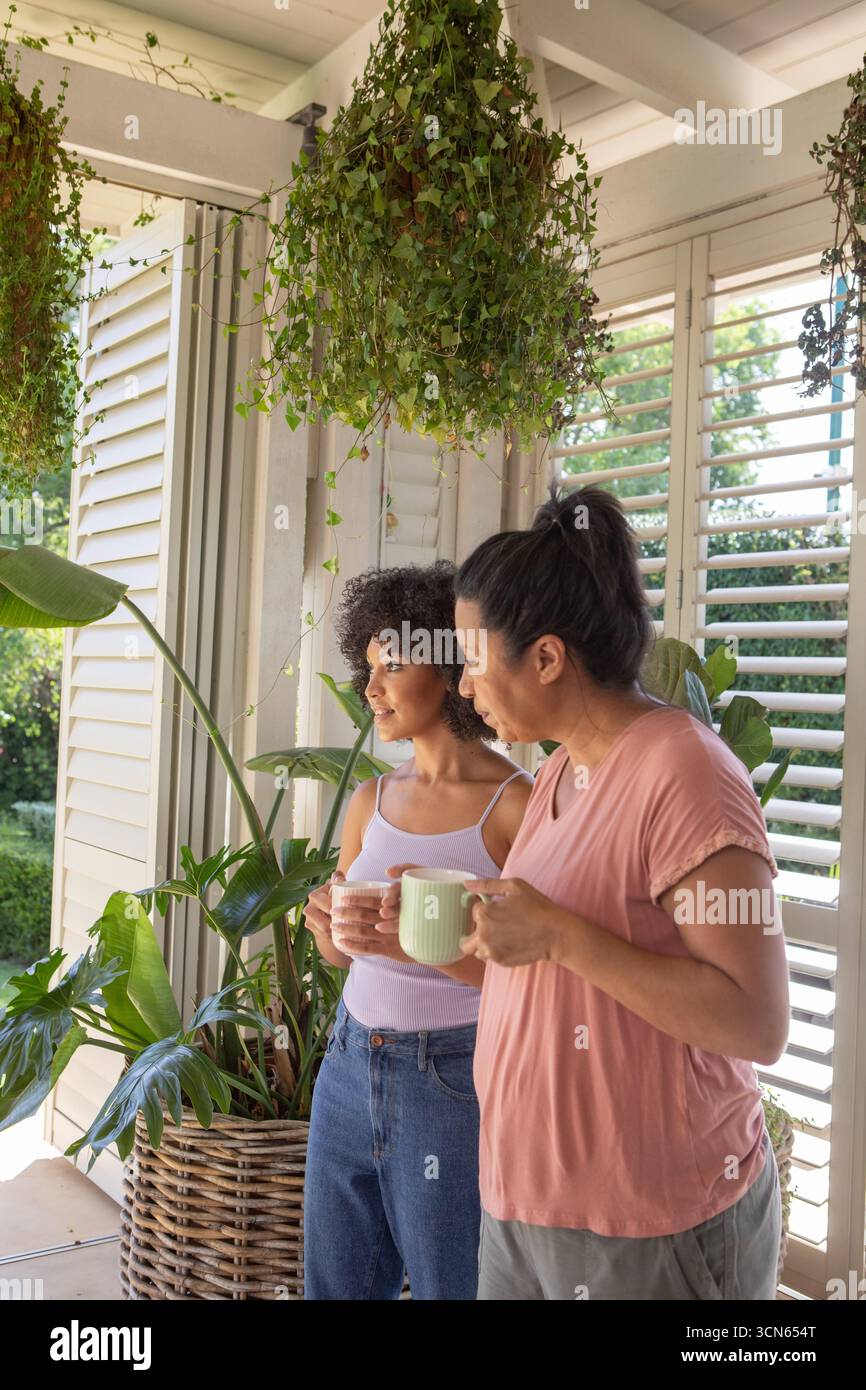 Mère et fille debout sur le porche par des volets tenant des tasses en céramique admirant la cour Banque D'Images