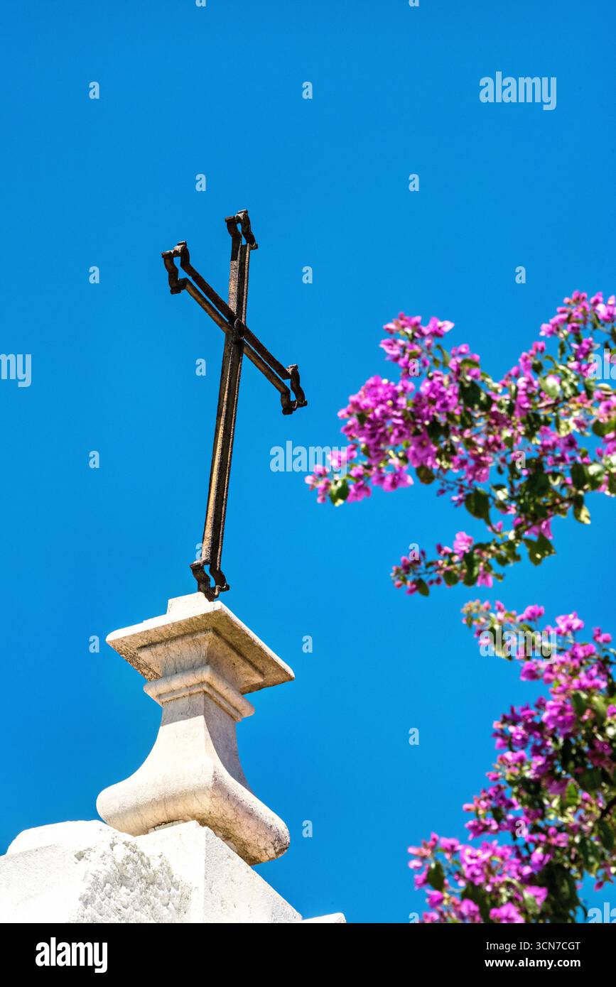 Croix d'église avec Bougainvillea Lisbonne Portugal // LISBONNE, Portugal — Une croix métallique altérée se dresse au sommet d'un piédestal de pierre contre un ciel bleu vif, avec des fleurs de bougainvillea rose vif fleurissant à sa droite. La croix, symbole du christianisme, est un spectacle commun dans toute Lisbonne, une ville avec une riche histoire religieuse. La bougainvillière, une vigne à fleurs originaire d'Amérique du Sud, prospère dans le climat méditerranéen de Lisbonne et est fréquemment vue orner les bâtiments et les murs. Cette image capture un détail de la beauté architecturale et naturelle de la ville, typique de ses paysages de rues pittoresques. Banque D'Images