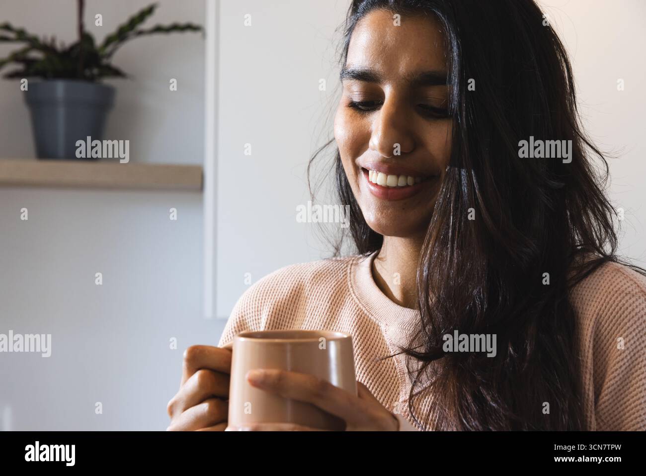 Femme asiatique tenant une tasse en céramique et souriant dans une cuisine à la maison lumineuse avec étagère en bois Banque D'Images