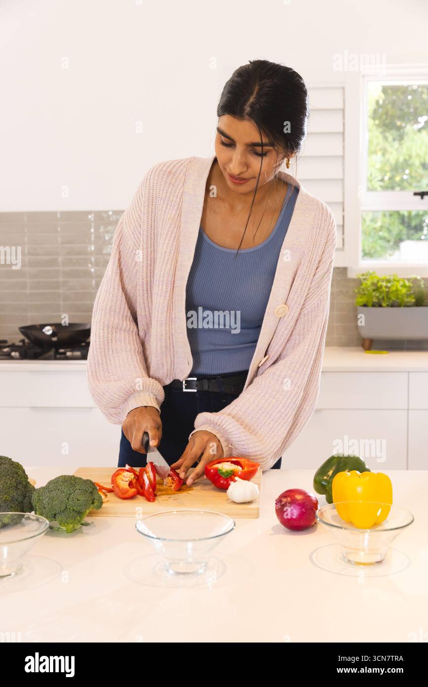 Femme indienne hachant le poivron rouge avec le couteau du chef sur la planche à découper à l'îlot de cuisine à la maison Banque D'Images