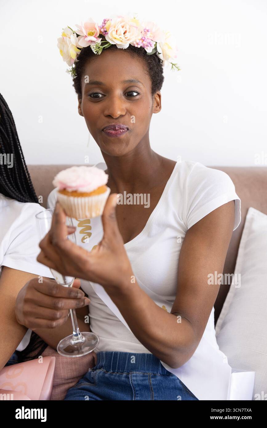 Amies afro-américaines assises sur le canapé portant une couronne, tenant un cupcake et une flûte à champagne Banque D'Images