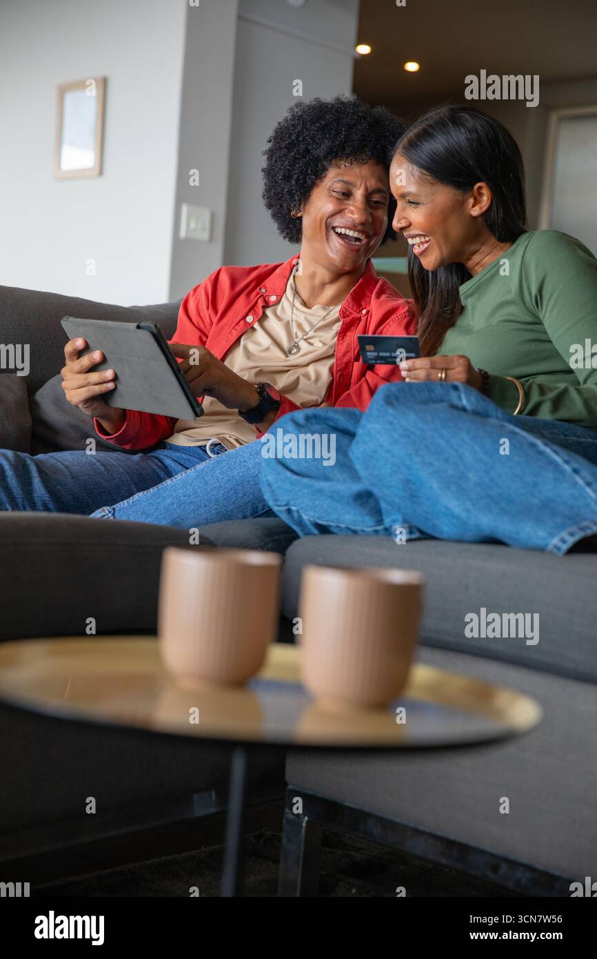 Diverses amies féminines appuyées ensemble sur un canapé gris à la maison, tablette de navigation, carte de crédit Banque D'Images Diverses amies féminines appuyées ensemble sur un canapé gris à la maison, tablette de navigation, carte de crédit Banque D'Images
