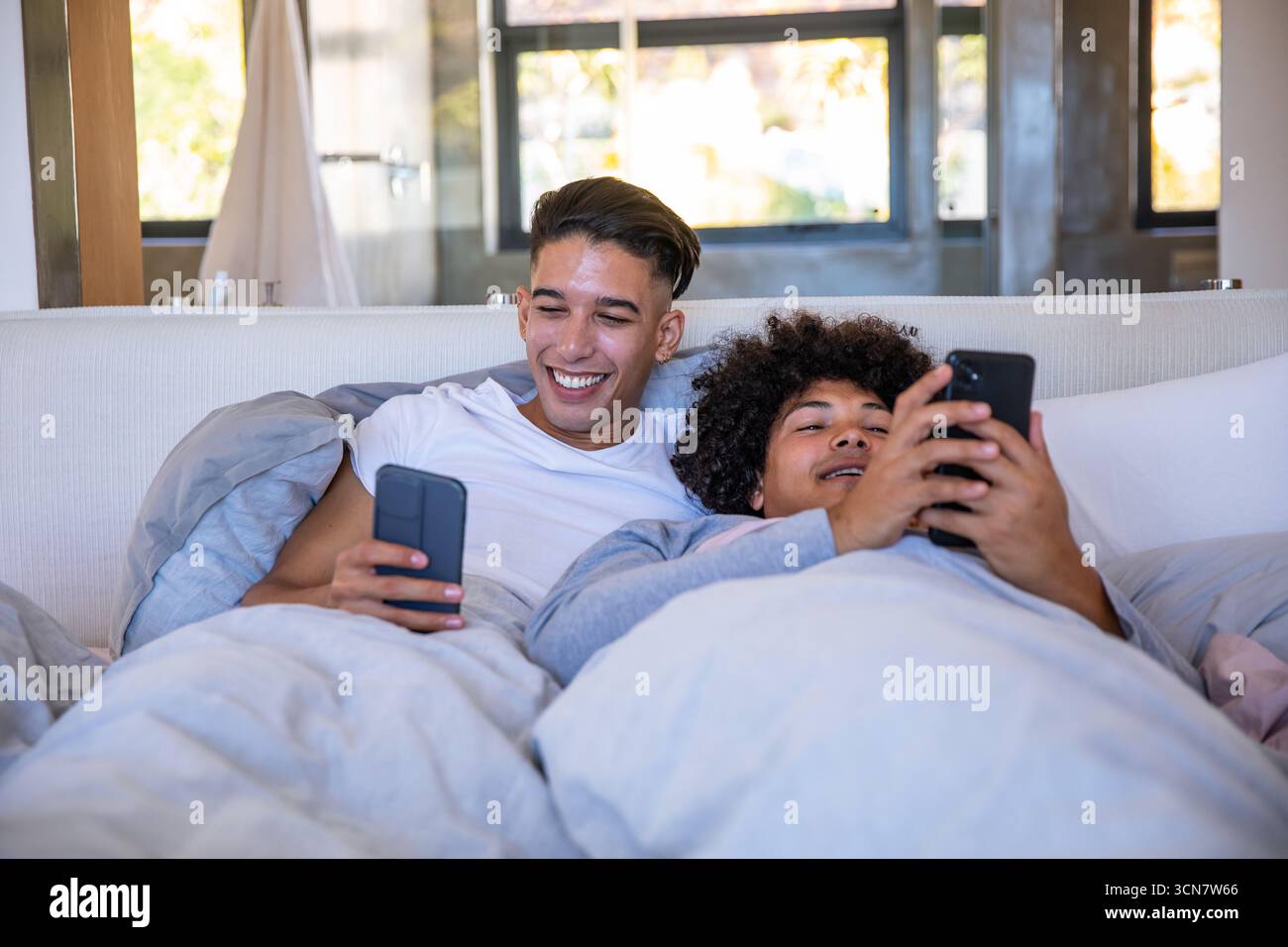 Divers partenaires masculins inclinés côte à côte sur le lit sous couette à l'aide de smartphones dans la chambre Banque D'Images