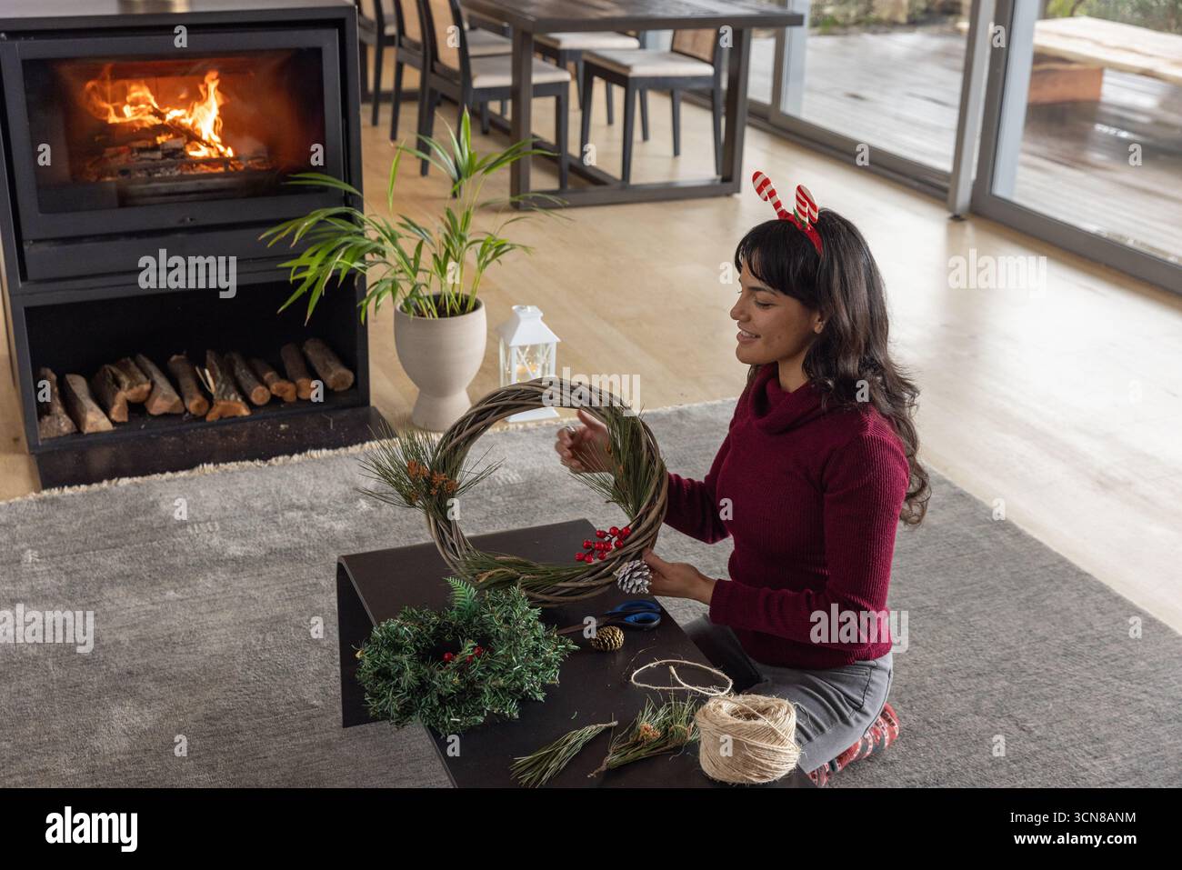 Femme asiatique assemblant une couronne de brindilles avec des pommes de pin, ficelle sur une table basse dans le salon Banque D'Images