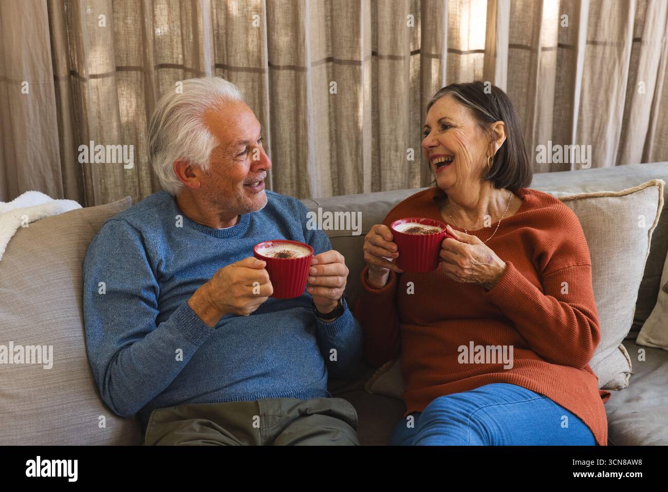 Couple senior tenant des tasses rouges et riant sur le canapé beige dans le salon partageant la pause café Banque D'Images
