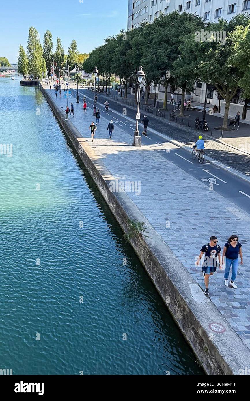 Coureurs, piétons et joggeurs profitent d'une journée beuatifiante le long du canal de l'Ourcq dans le 19ème arrondissement de Paris le 7 septembre 2025. Un large canal urbain ou une rivière coule entre deux promenades bordées d'arbres dans un cadre urbain. A droite, une large passerelle en pierre est bordée par une balustrade et des piétons ; à gauche, un bas bord de pierre mène à l'eau. Au loin, des arbres et un groupe de bâtiments de faible hauteur et une grande structure blanche moderne s'élèvent contre un ciel bleu clair. La scène semble calme et ensoleillée, avec la lumière qui se reflète sur l'eau. (Photo de Samuel Rigelhaupt/Sipa USA) Banque D'Images