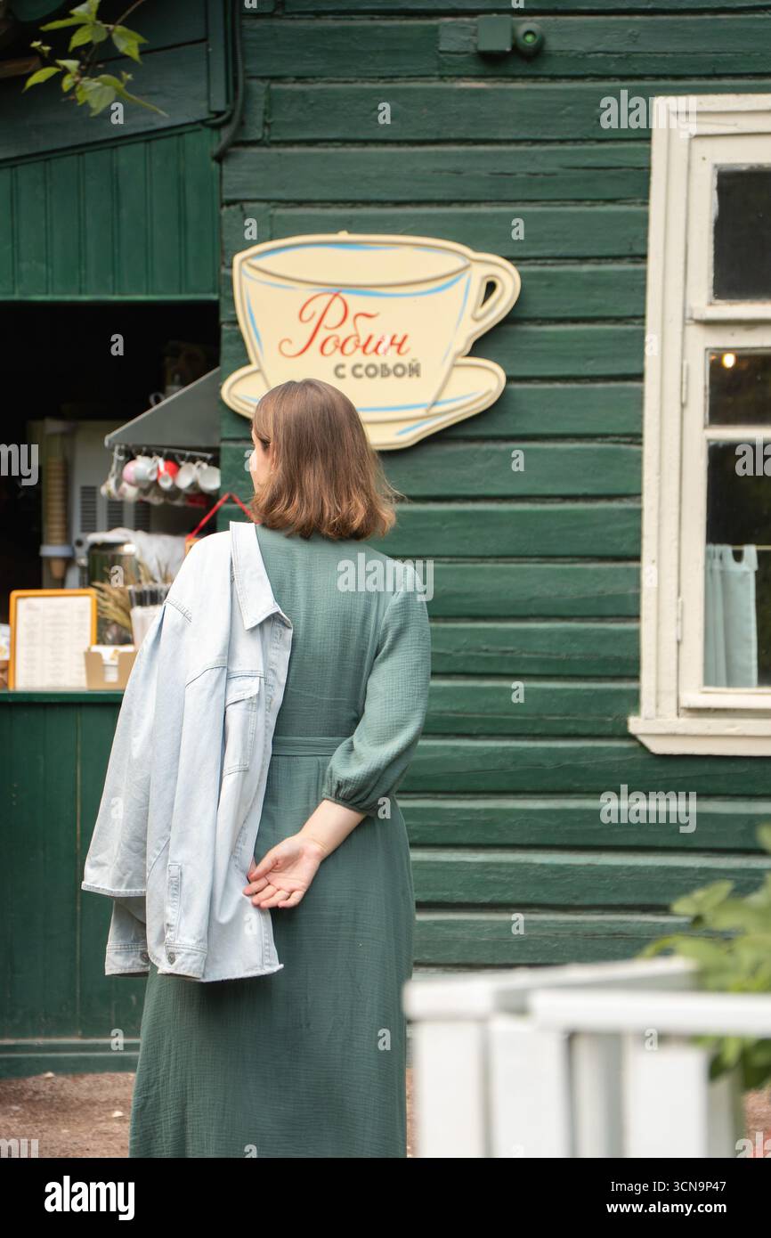 Peredelkino, Russie - 9 août 2025 : femme dans une robe verte tenant une veste en denim se tient près du café en bois Robin avec un signe en forme de tasse Banque D'Images