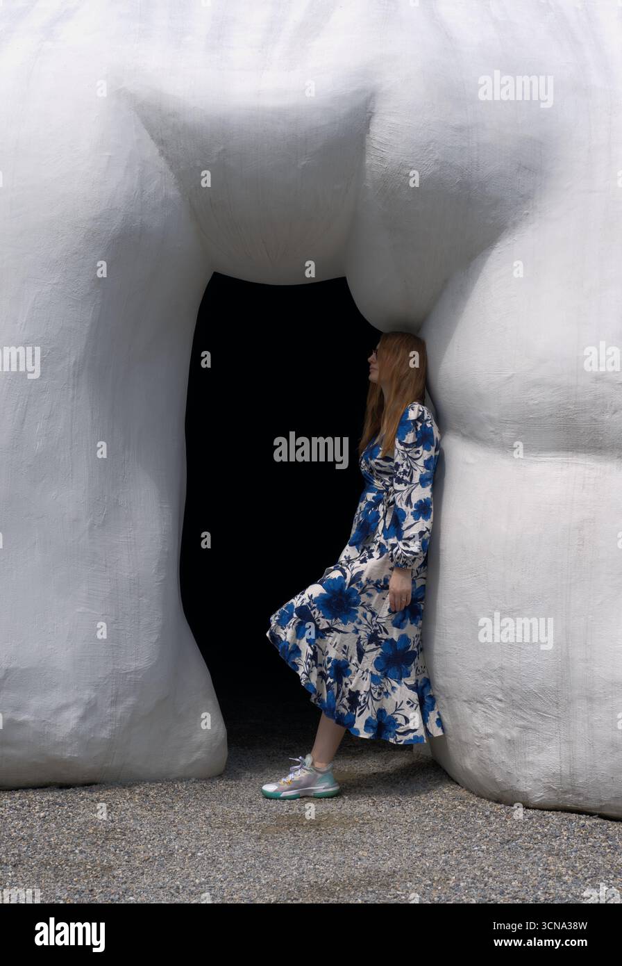 Une personne aux cheveux longs vêtue d'une robe florale bleue et blanche marche vers une entrée sombre d'un bâtiment encadrée par deux grandes architectures blanches de forme organique Banque D'Images