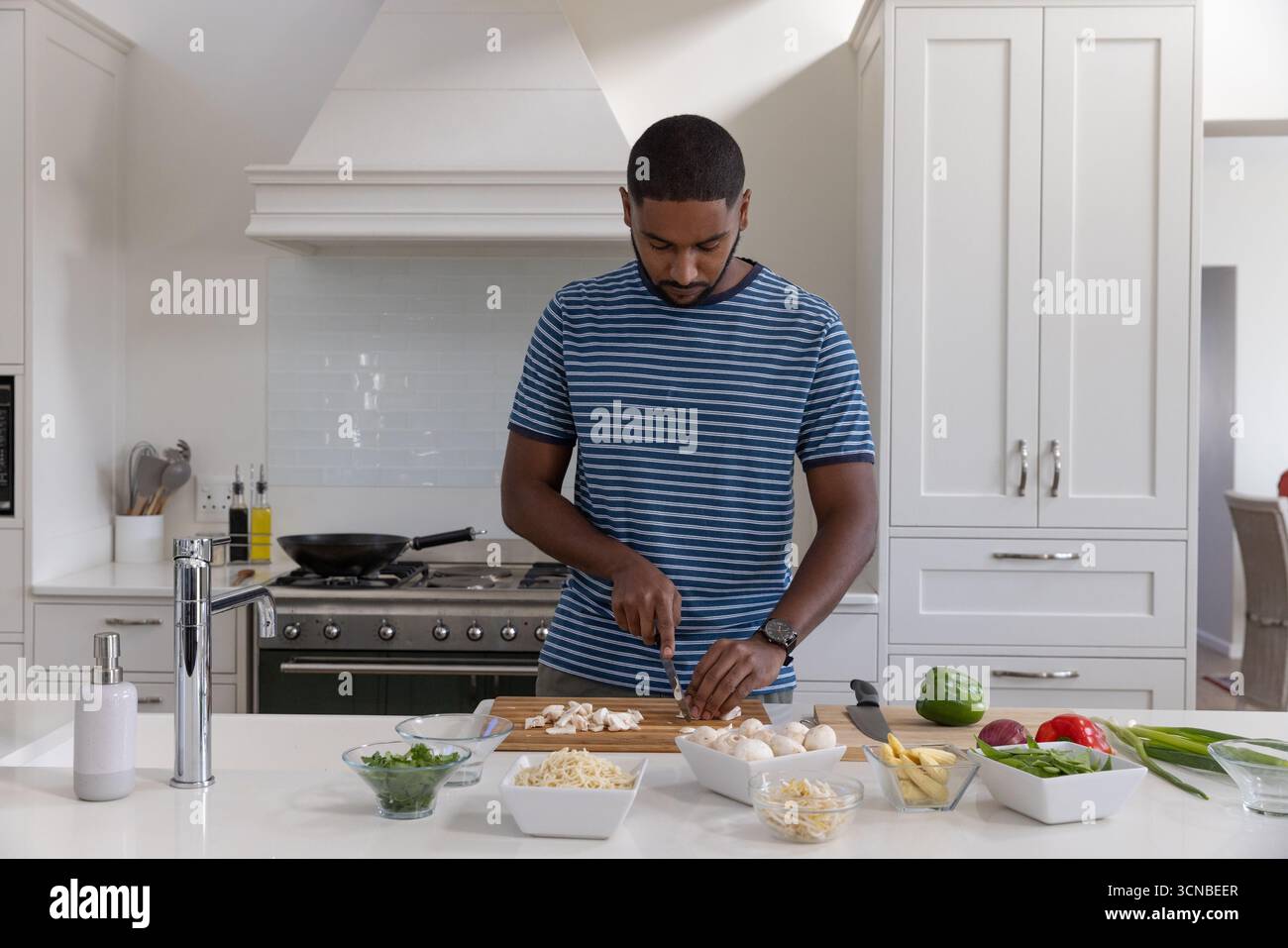 Homme afro-américain tranchant des champignons dans la cuisine à la maison à l'île avec couteau de chef et bols Banque D'Images
