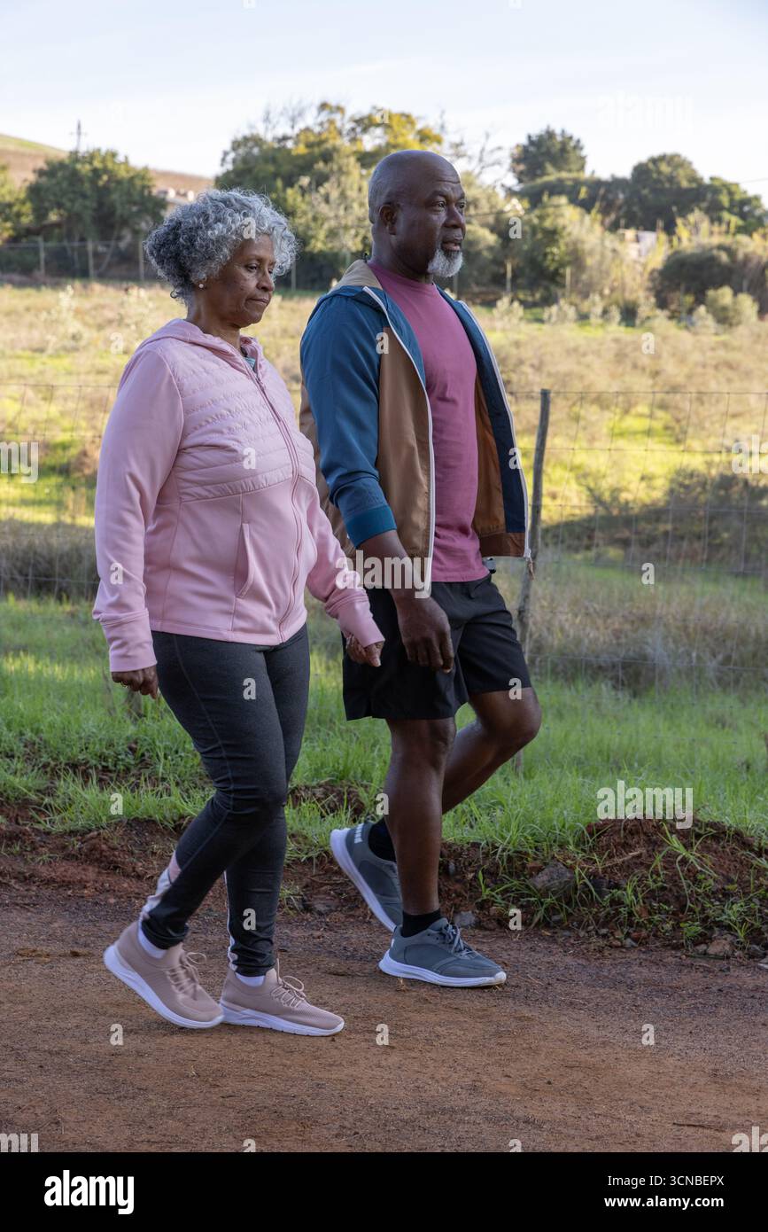 Couple senior afro-américain marchant sur le sentier de terre à côté de la clôture de maillons de chaîne portant des sweats à capuche Banque D'Images