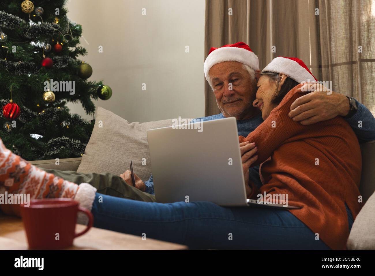 Couple senior embrassant portant des chapeaux de Père Noël sur le canapé dans le salon avec ordinateur portable et arbre de Noël Banque D'Images