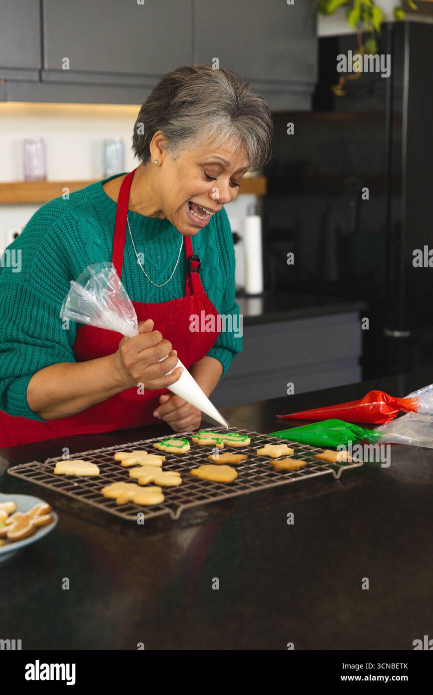 Femme senior dans le tablier rouge au comptoir de cuisine en utilisant le sac de tuyauterie décorant les biscuits avec le glaçage blanc Banque D'Images