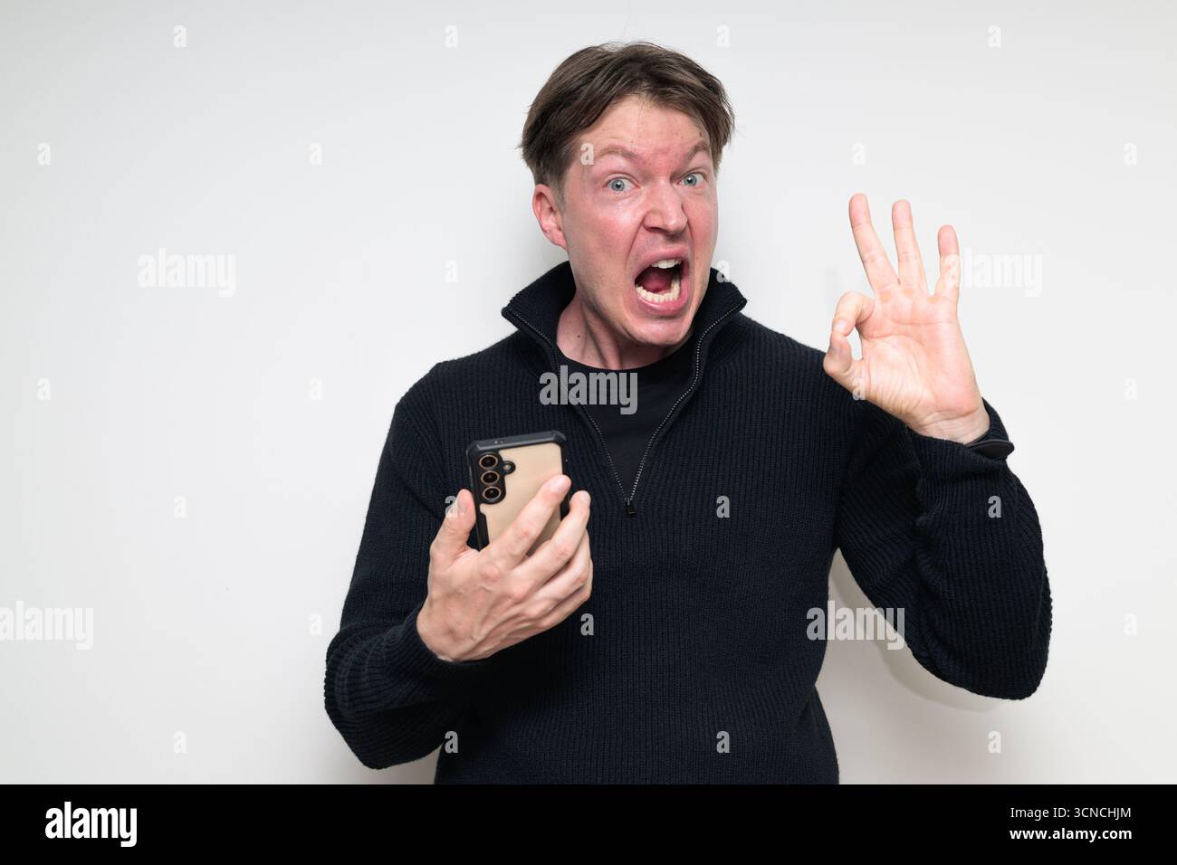 Heureux homme adulte scandinave moyen à l'aide du téléphone sur fond blanc faisant le geste de signe OK Banque D'Images