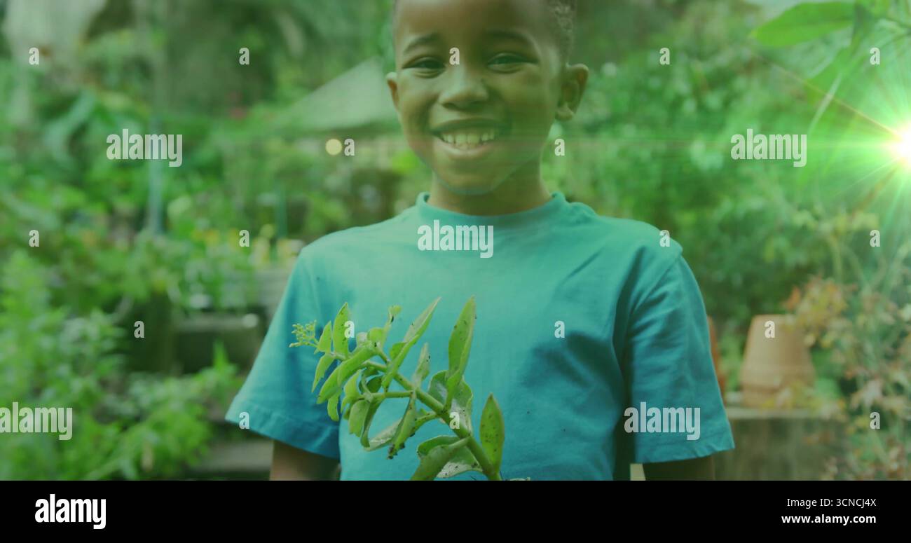 Garçon afro-américain souriant tenant une branche de plante feuillue dans la serre, avec des bancs et des pots de plantes Banque D'Images
