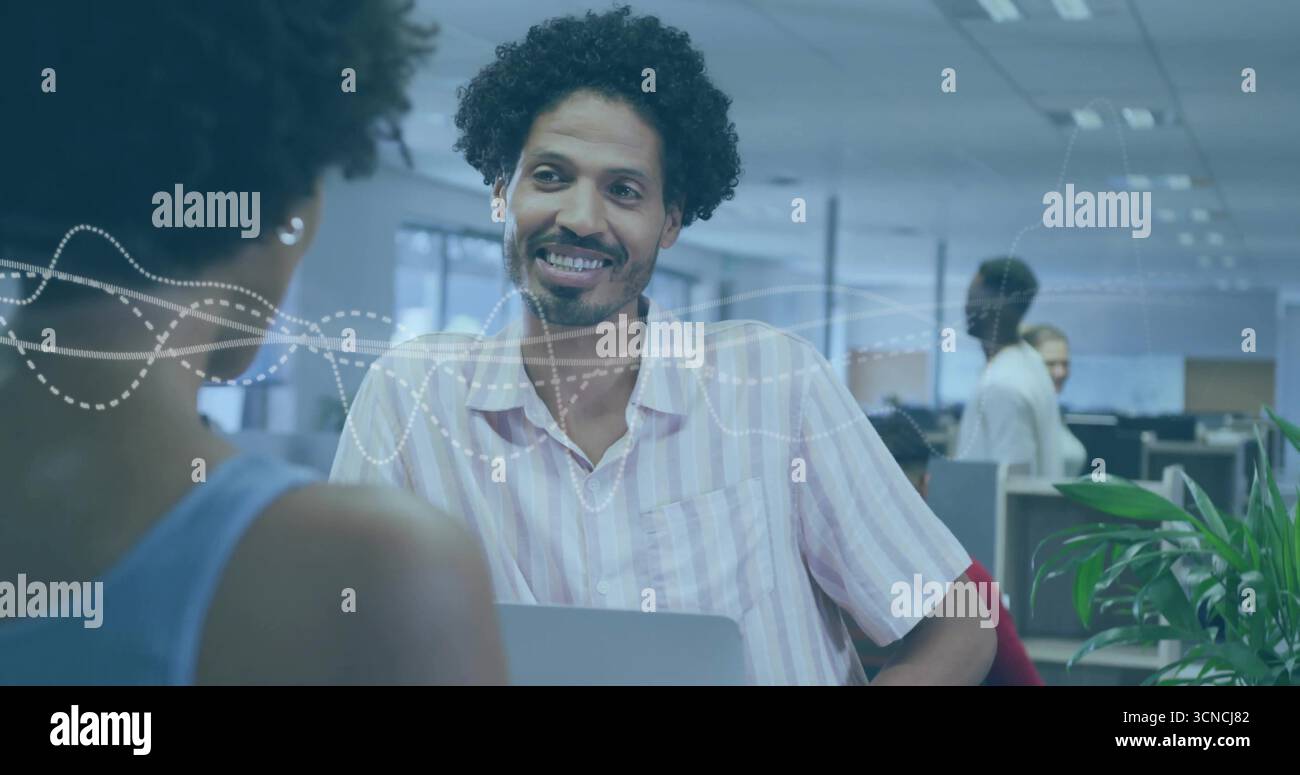 Homme souriant en chemise rayée discutant avec un collègue au bureau ouvert, avec ordinateur portable et moniteurs Banque D'Images