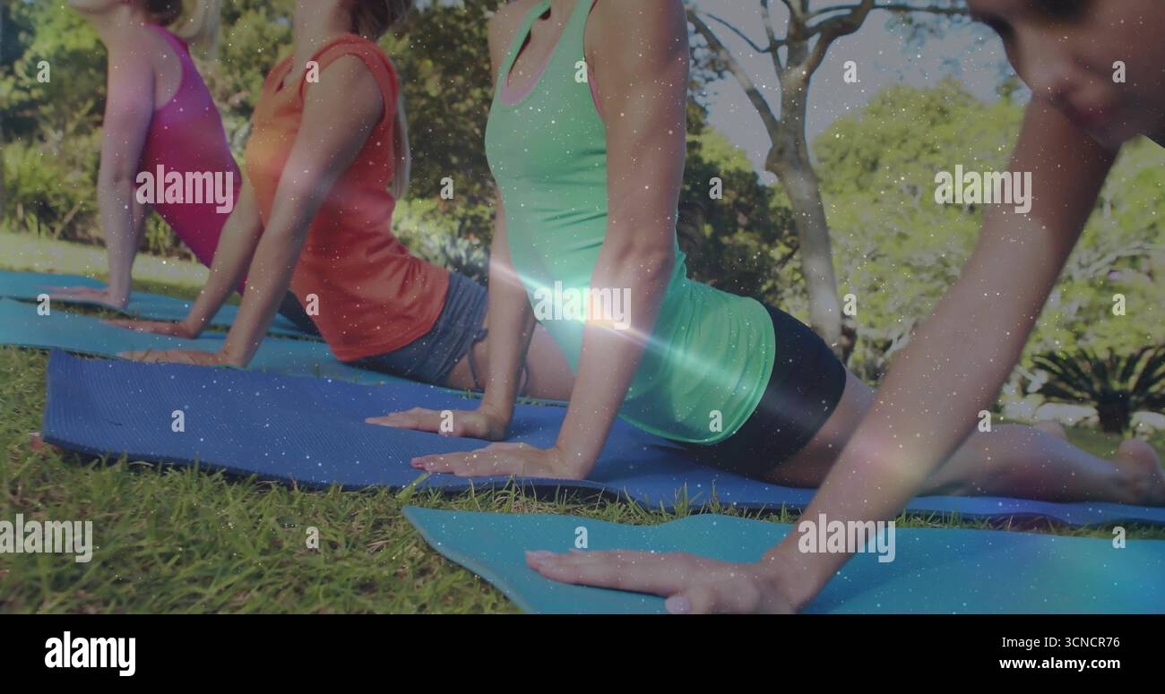 Se penchant vers le dos, quatre amis pressant les mains dans des tapis de yoga dans un parc herbeux, avec des vêtements athlétiques brillants Banque D'Images