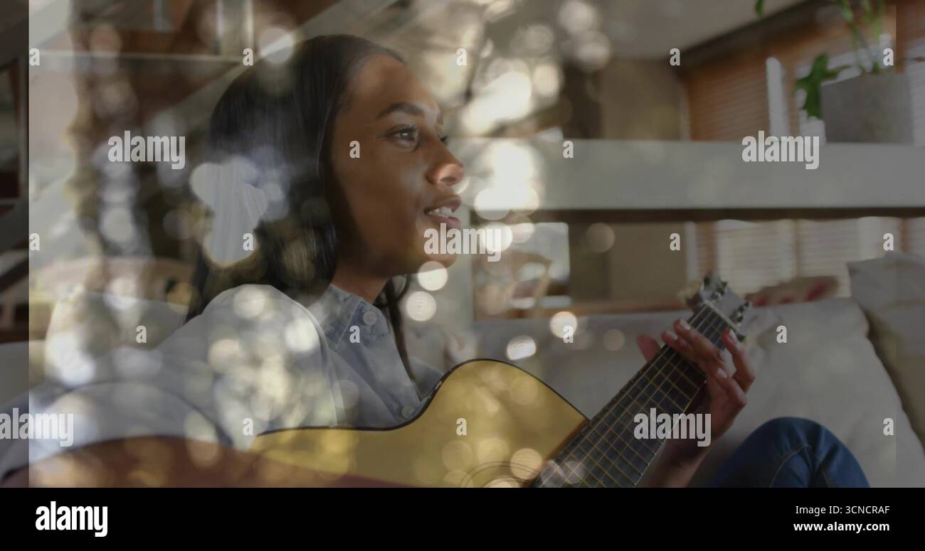 Musicien de guitare acoustique strumming portant une tenue en denim sur le canapé dans le salon, avec des lumières bokeh Banque D'Images