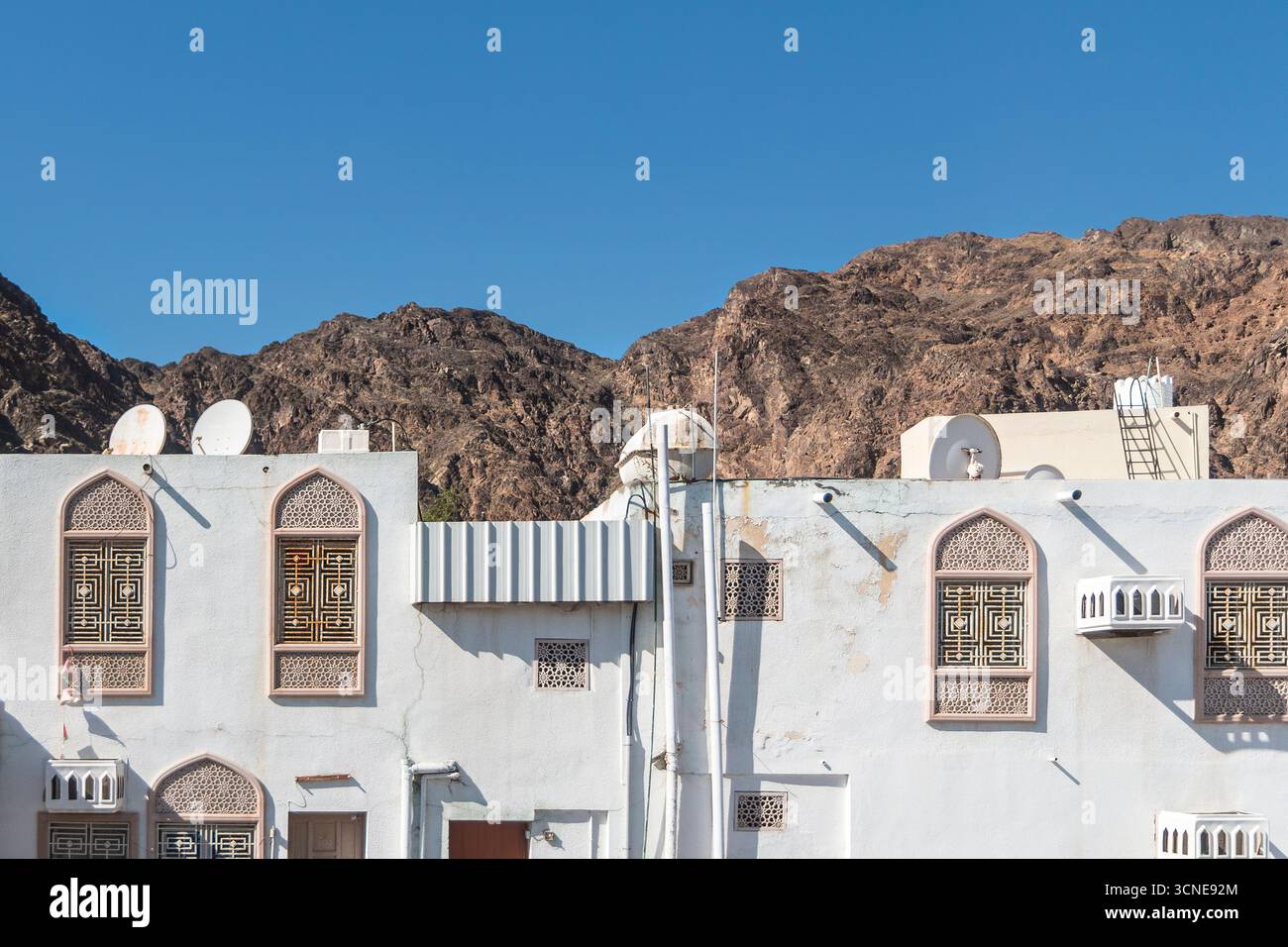 Oman - Muscat - vieille ville - façades blanches ensoleillées avec fenêtres ornées sous des collines brunes dentelées. Banque D'Images