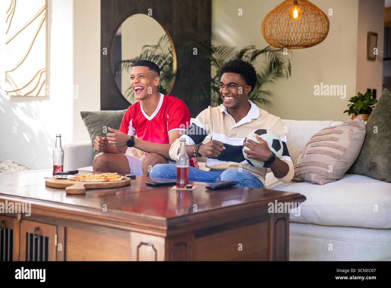 Divers amis masculins riant tout en tenant un ballon de football et en mangeant de la pizza sur le canapé du salon Banque D'Images