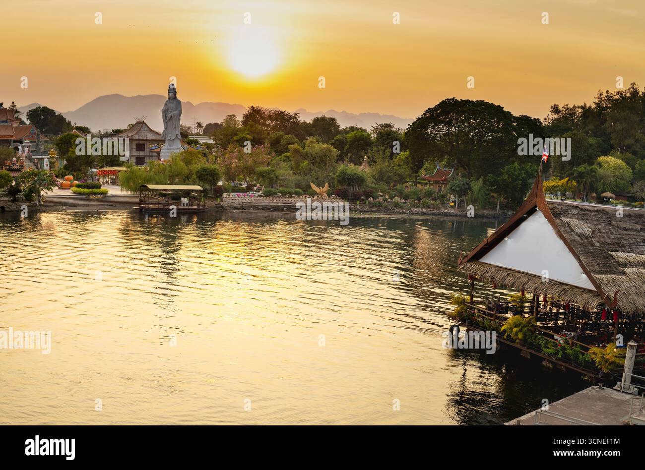 Le soleil se couche derrière une statue de temple bouddhiste, vers des montagnes lointaines, lumière chaude dorée reflétant de l'eau de la rivière, à l'attrac touristique populaire Banque D'Images