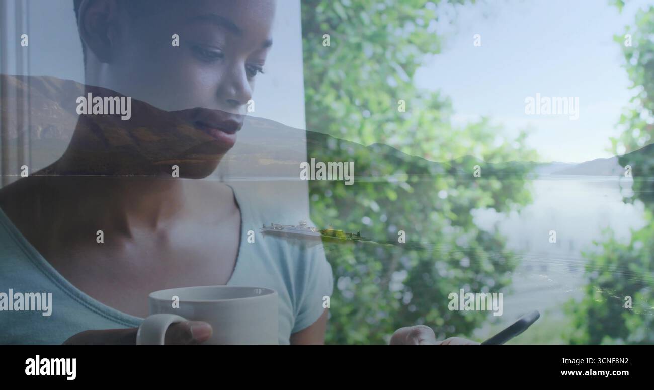 Femme debout portant un haut sarcelle tenant une tasse blanche et un smartphone par la fenêtre regardant le lac, espace de copie Banque D'Images
