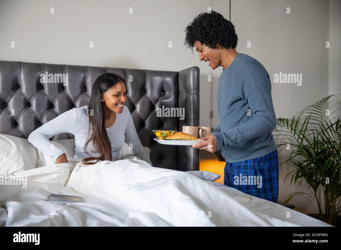 Couple indien et afro-américain assis dans le lit tenant plateau de petit déjeuner, tasses à café, croissant Banque D'Images Couple indien et afro-américain assis dans le lit tenant plateau de petit déjeuner, tasses à café, croissant Banque D'Images