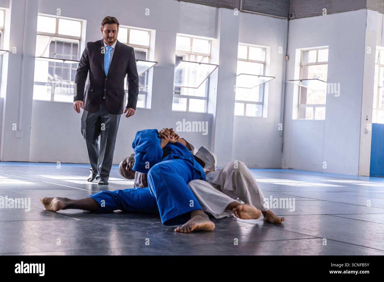 Divers concurrents en bleu blanc gis grappling sur des tapis dans la salle martiale avec l'observation des arbitres Banque D'Images