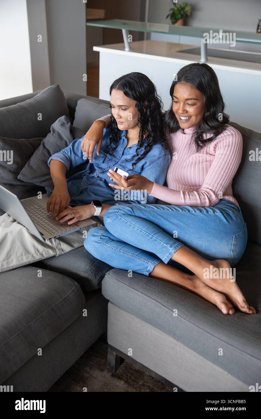 Amies afro-américaines assises sur le canapé à la maison, à l'aide d'un smartphone, pointant vers l'ordinateur portable Banque D'Images Amies afro-américaines assises sur le canapé à la maison, à l'aide d'un smartphone, pointant vers l'ordinateur portable Banque D'Images