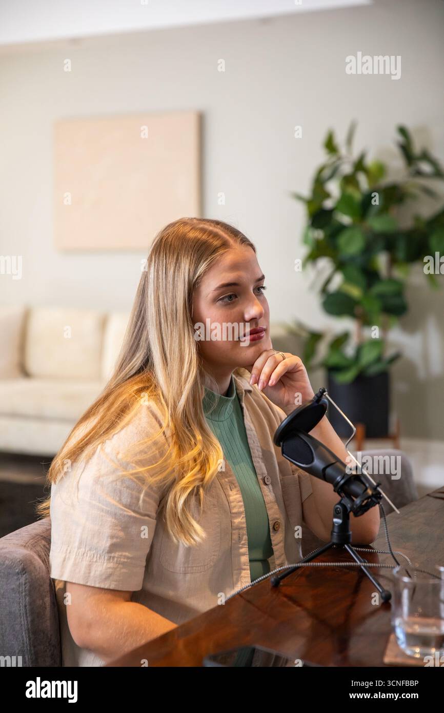 Femme créatrice de contenu enregistrant podcast à table dans le salon avec microphone, verre d'eau Banque D'Images