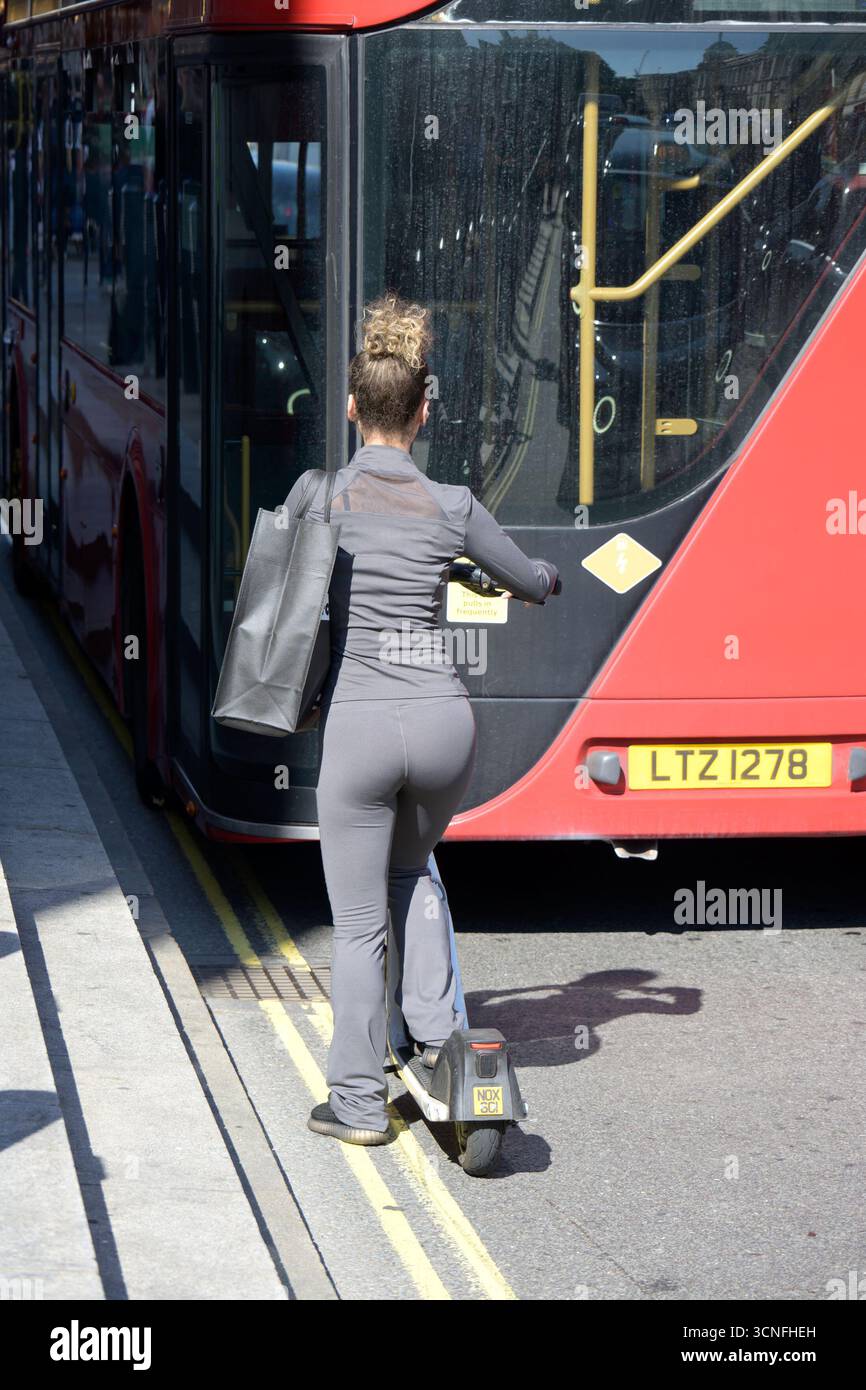 Londres, Royaume-Uni. Jeune femme à la mode chevauchant un scooter électrique derrière un bus Banque D'Images