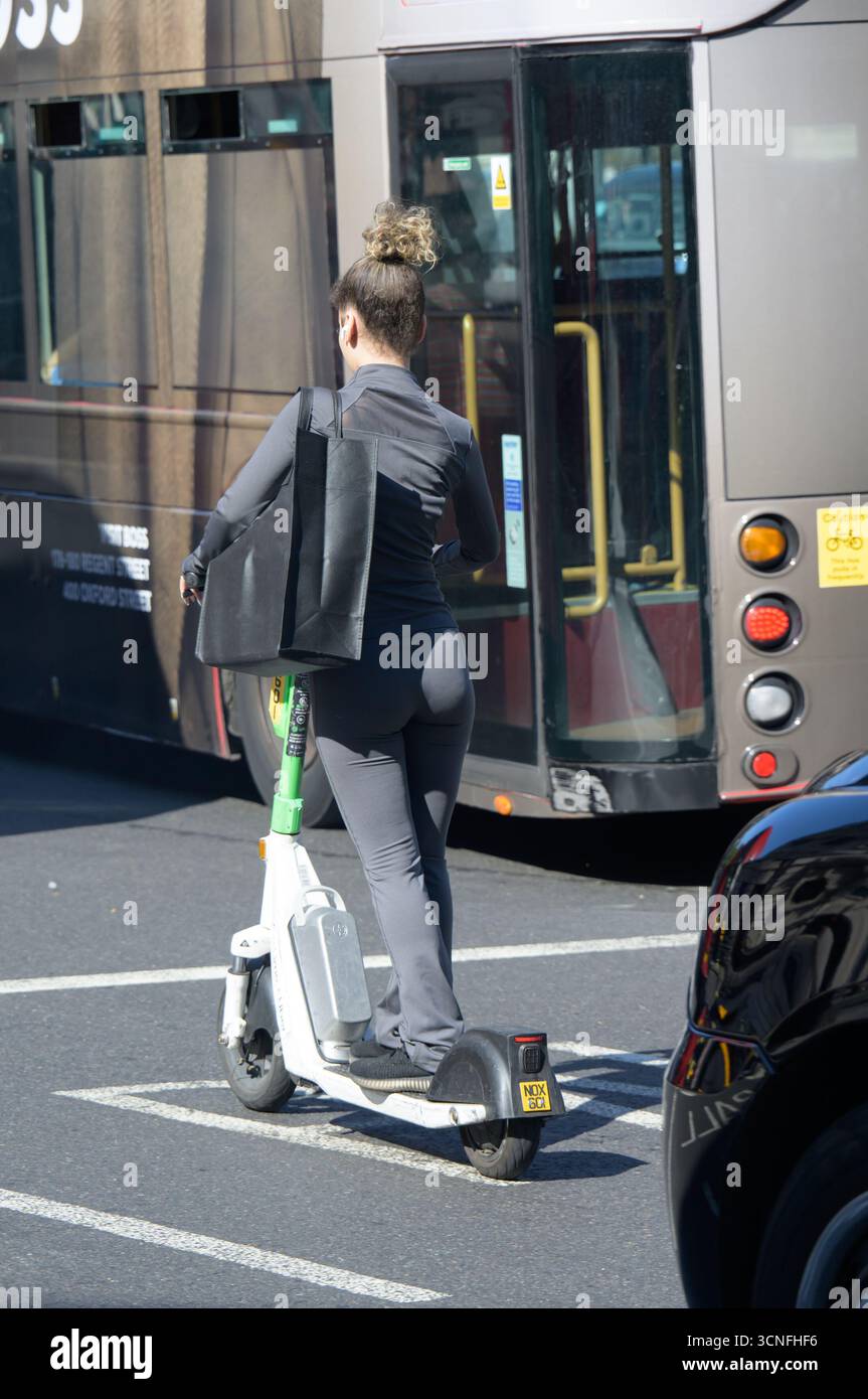 Londres, Royaume-Uni. Jeune femme à la mode ridng un scooter électrique derrière un bus Banque D'Images