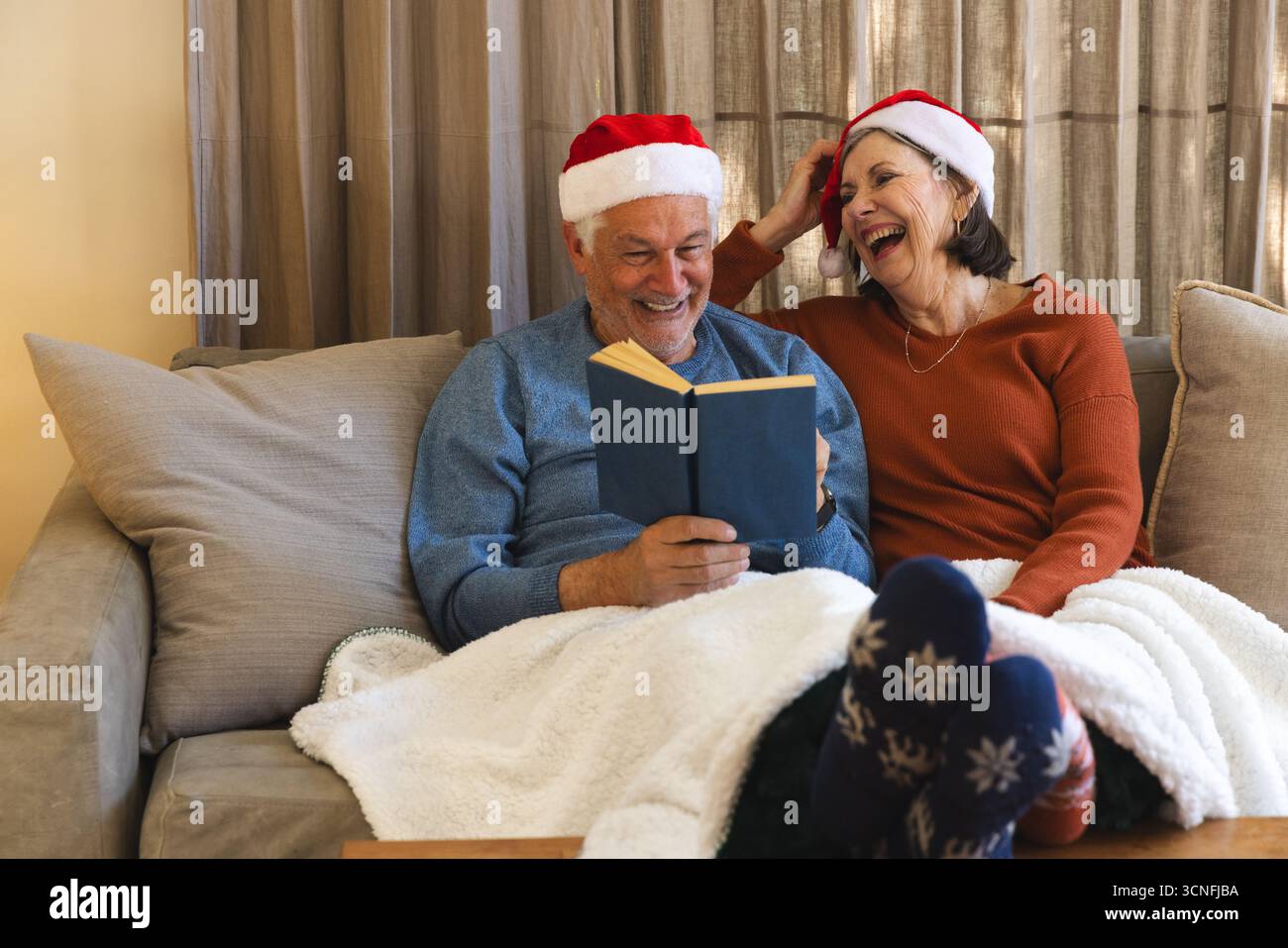 Couple senior portant des chapeaux de Père Noël assis sur un canapé sous une couverture en polaire blanche, lisant un livre à couverture rigide Banque D'Images