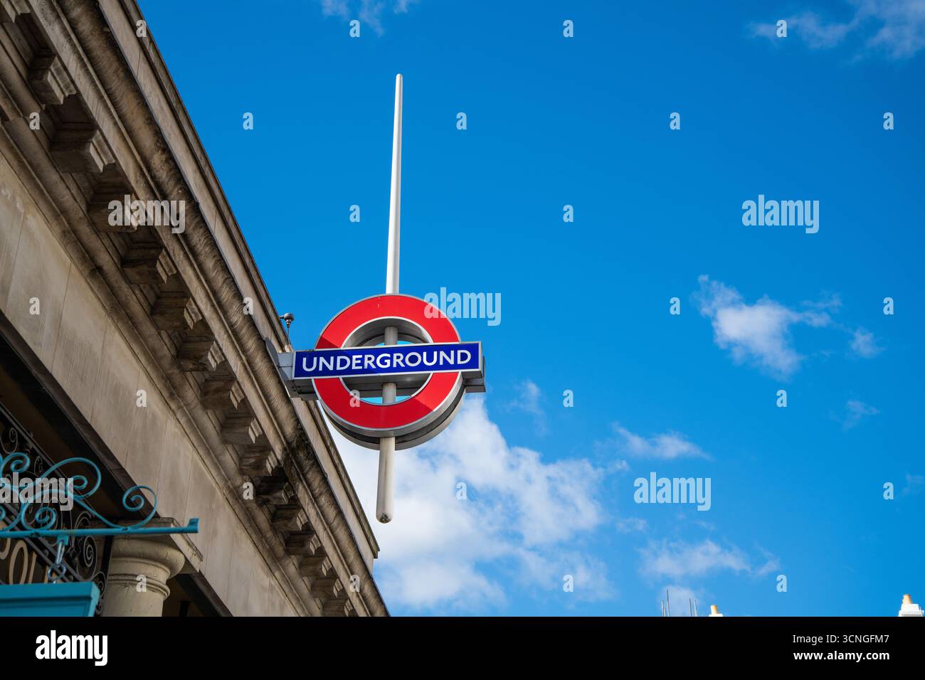 Gros plan de l'emblématique panneau rouge et bleu du métro londonien sur la façade d'un bâtiment, face à un ciel bleu clair. Banque D'Images