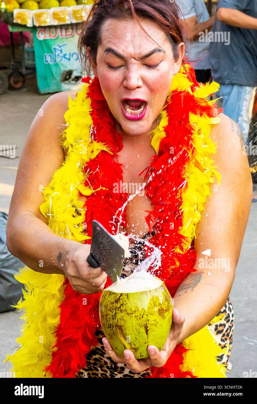 Kanchanaburi, Thaïlande-janvier 31 2025 : une femme transgenre extravagante, habillée de couleurs vives, crie fort tout en ouvrant rapidement les noix de coco, à vendre t Banque D'Images