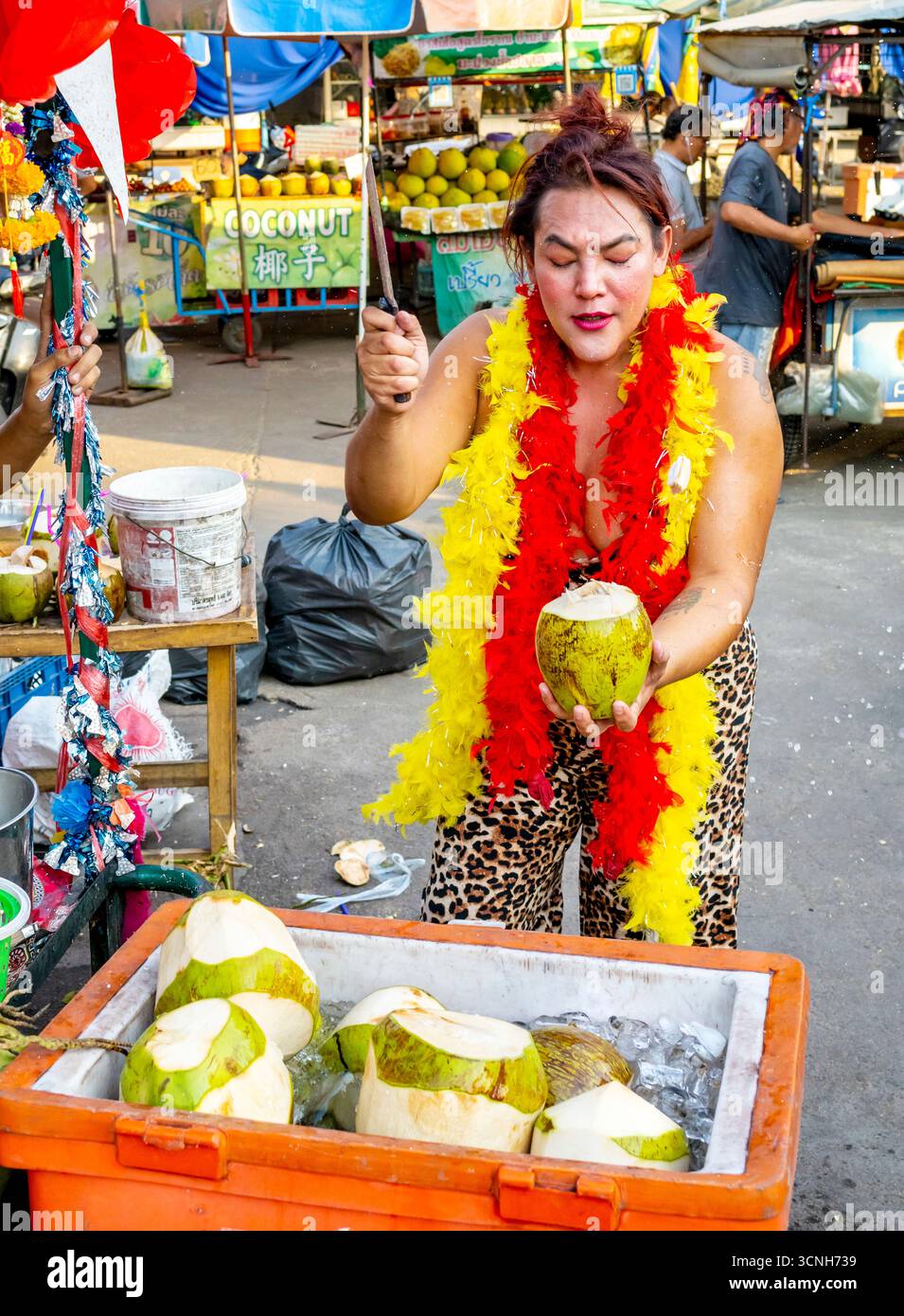Kanchanaburi, Thaïlande-janvier 31 2025 : une femme transgenre extravagante, habillée de couleurs vives, crie fort tout en ouvrant rapidement les noix de coco, à vendre t Banque D'Images