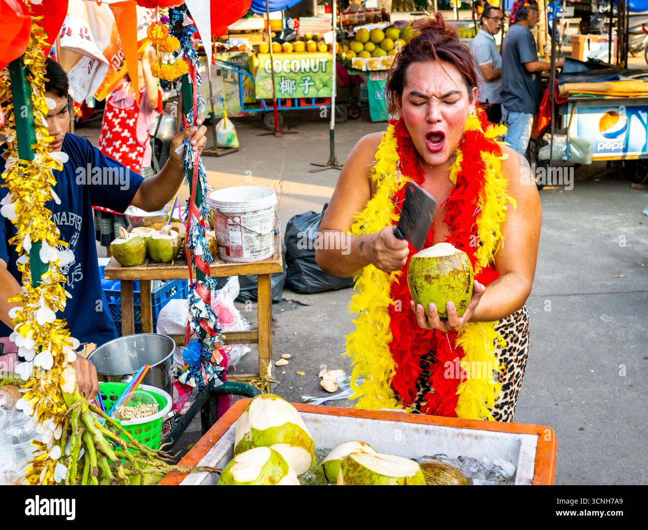 Kanchanaburi, Thaïlande-janvier 31 2025 : une femme transgenre extravagante, habillée de couleurs vives, crie fort tout en ouvrant rapidement les noix de coco, à vendre t Banque D'Images