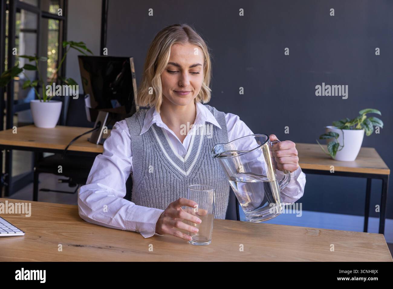 Travailleuse de bureau versant l'eau du pichet dans le verre au bureau avec moniteur et plantes Banque D'Images