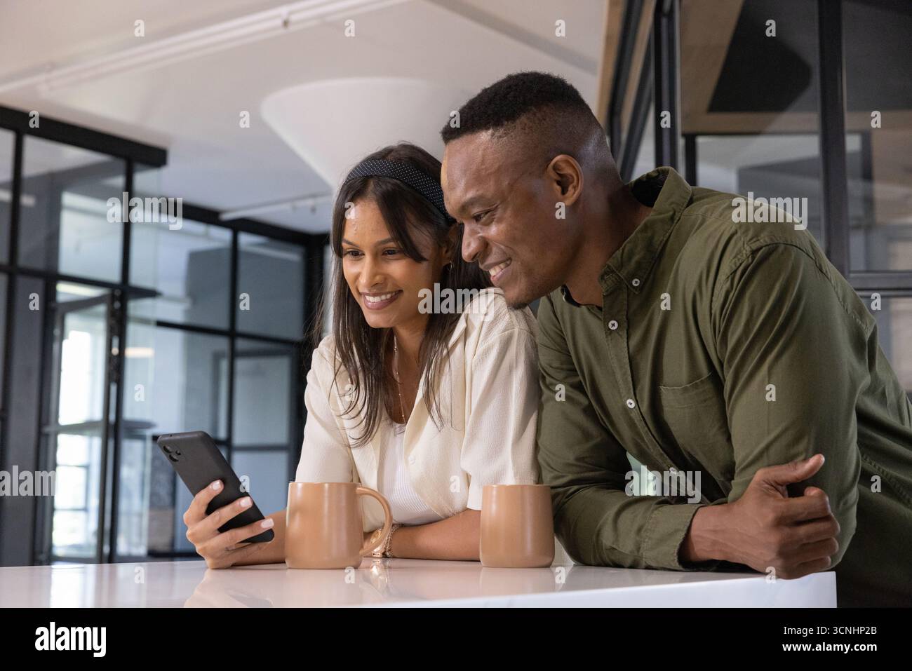 Divers collègues s'appuyant sur le comptoir blanc dans la salle de pause partageant un smartphone près de deux tasses bronzées Banque D'Images