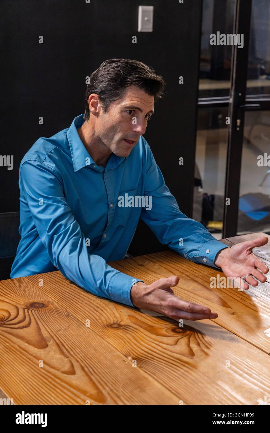 Homme en chemise bleue penché en avant avec les paumes vers le haut à la table de réunion à côté de la cloison de verre Banque D'Images