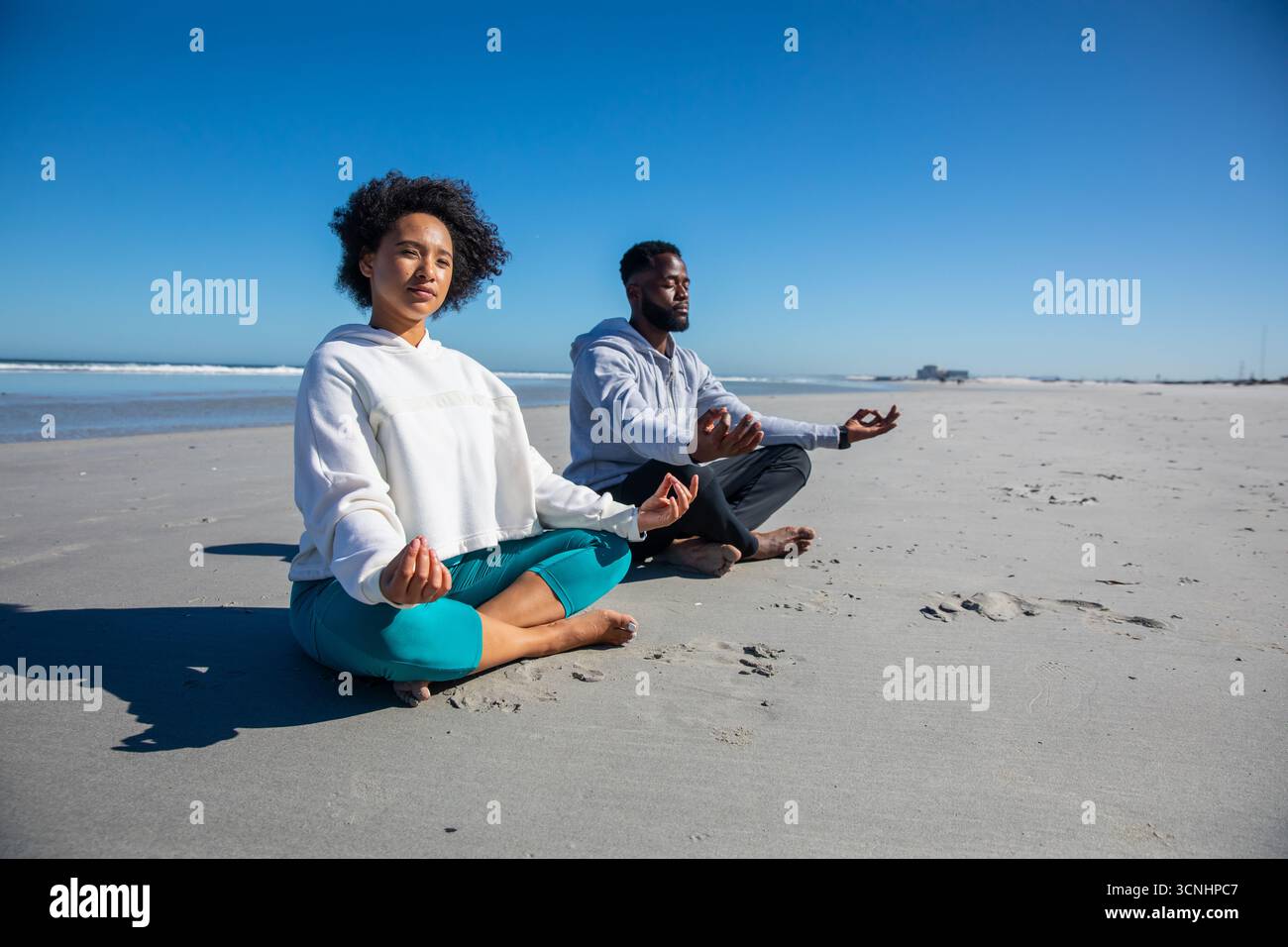Couple afro-américain méditant en lotus sur la plage près du bord de l'eau en sweats à capuche et leggings Banque D'Images