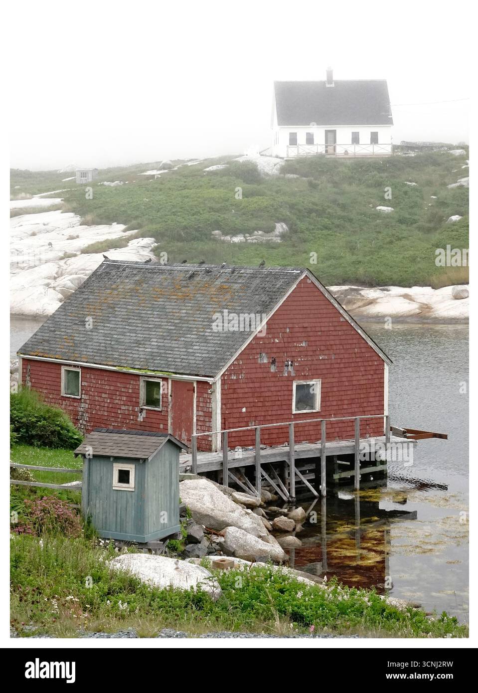 Une scène côtière sereine à Peggy's Cove, en Nouvelle-Écosse, capture un hangar à bateaux rouge niché au bord de l'eau, entouré de brouillard. De vieilles maisons parsèment le paysage Banque D'Images