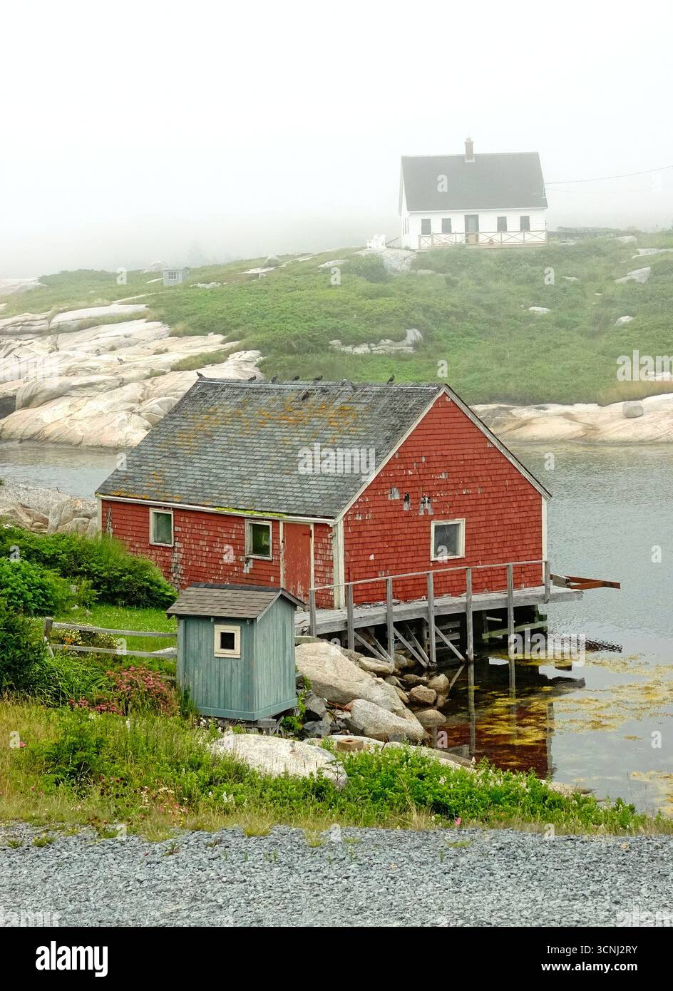 Une scène côtière sereine à Peggy's Cove, en Nouvelle-Écosse, capture un hangar à bateaux rouge niché au bord de l'eau, entouré de brouillard. De vieilles maisons parsèment le paysage Banque D'Images