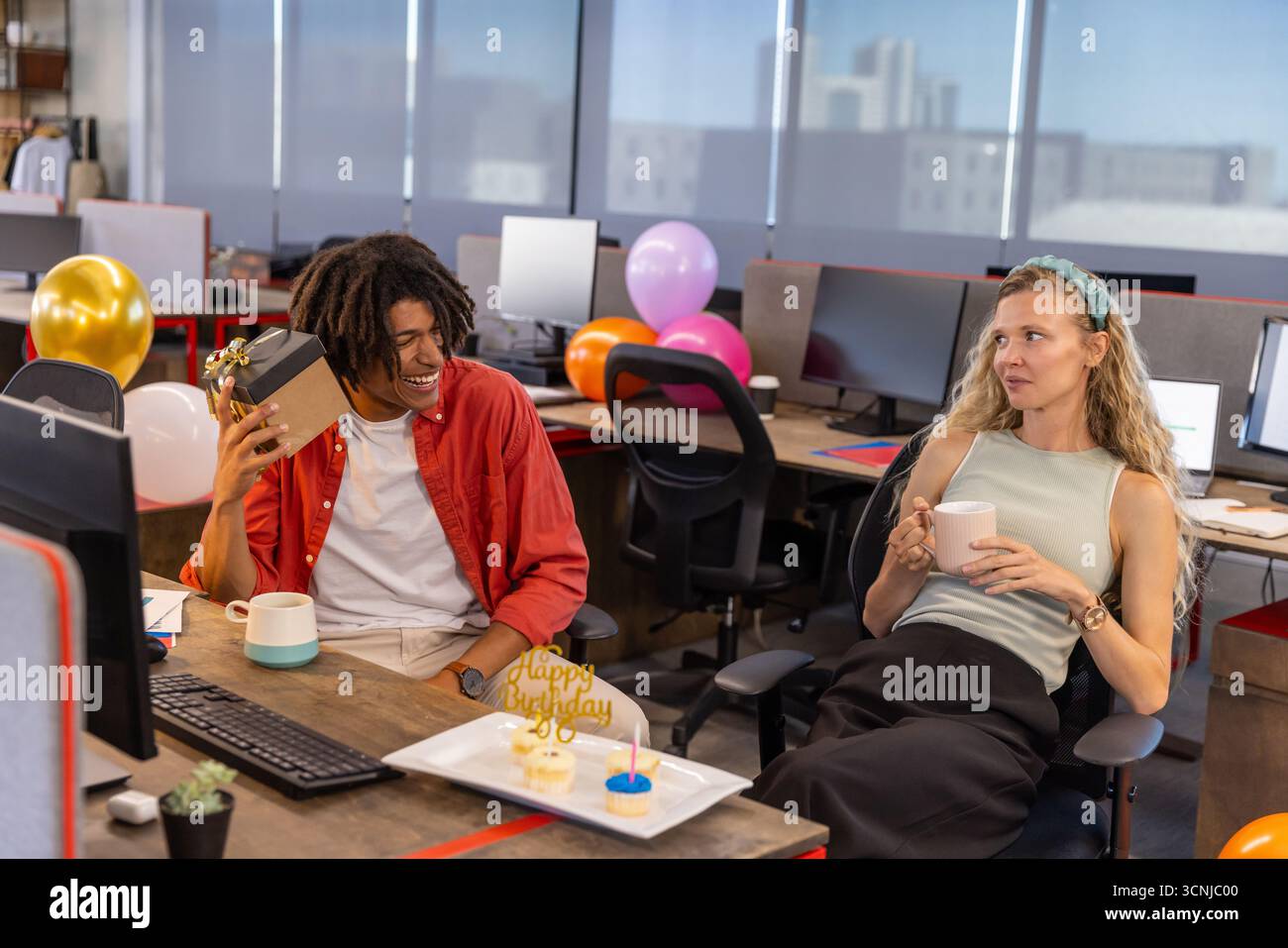 Divers collègues tenant un cadeau emballé et partageant des cupcakes au bureau décoré avec des ballons Banque D'Images