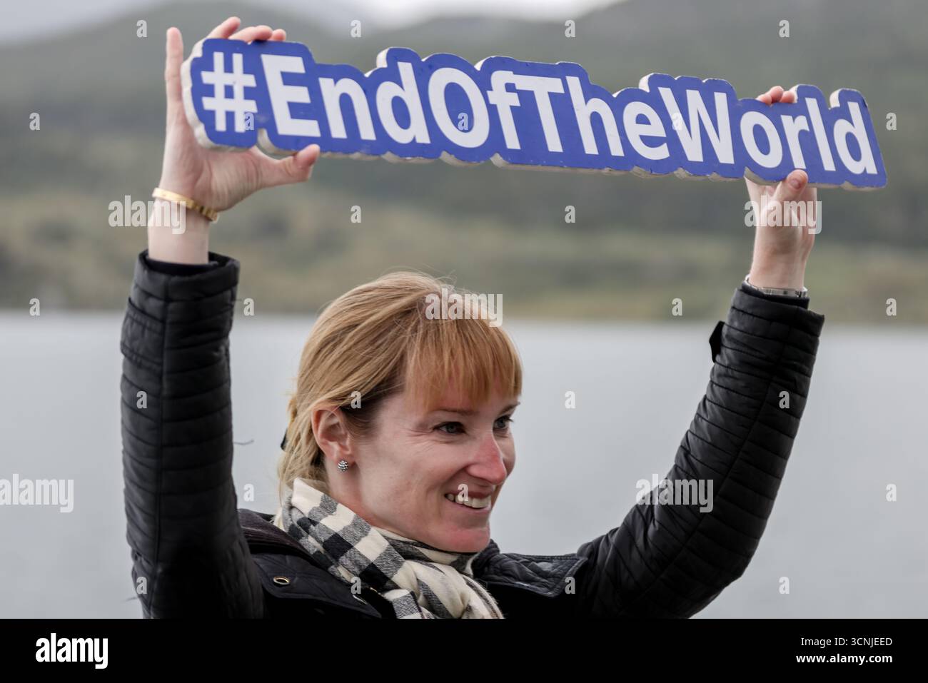 Un touriste souriant tient un panneau « #EndOfTheWorld » près du canal Beagle, célébrant le statut d’Ushuaia comme la ville la plus méridionale du monde. Banque D'Images