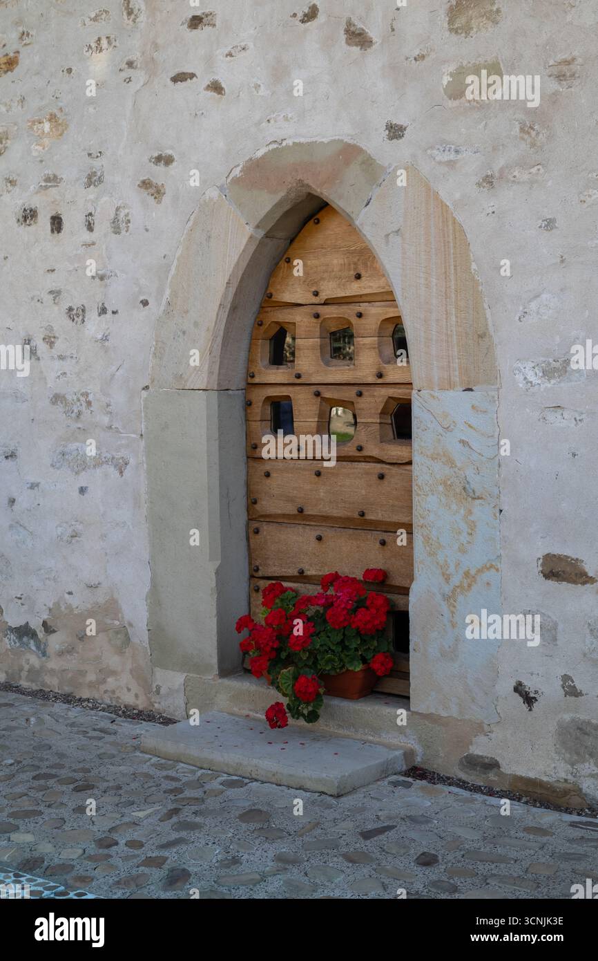 Une porte en bois voûtée ornée de géraniums rouges contre un ancien mur de pierre, créant une scène de charme européen rustique et de beauté intemporelle. Banque D'Images